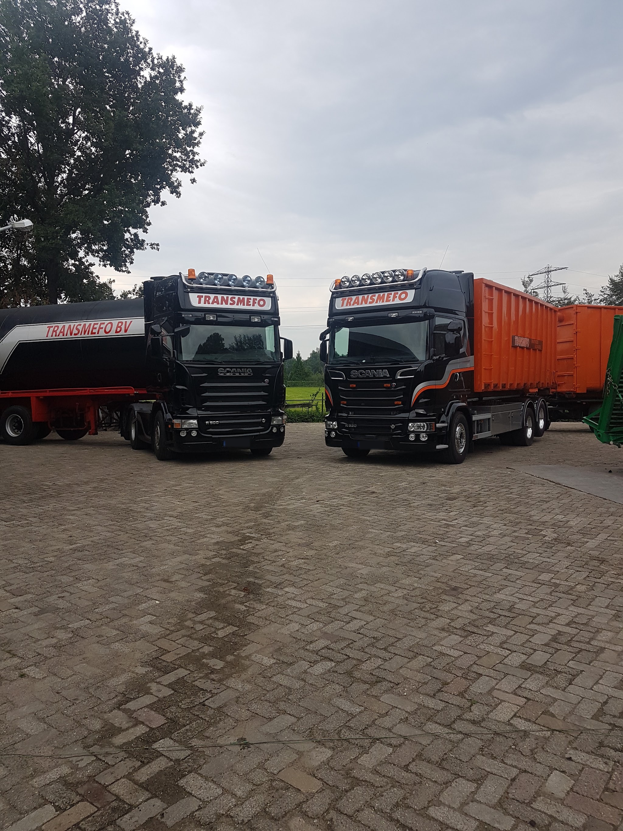 Twee zwarte Scania-vrachtwagens TRANSMEFO BV met containers naast elkaar op een klinkererf