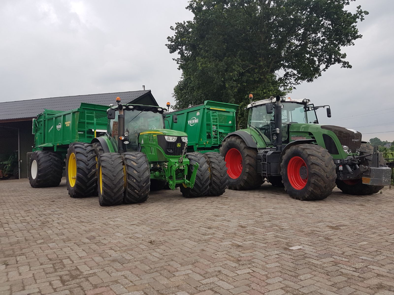 Twee trekkers, een John Deere en een Fendt, met groene aanhangers op een klinkererf bij een schuur