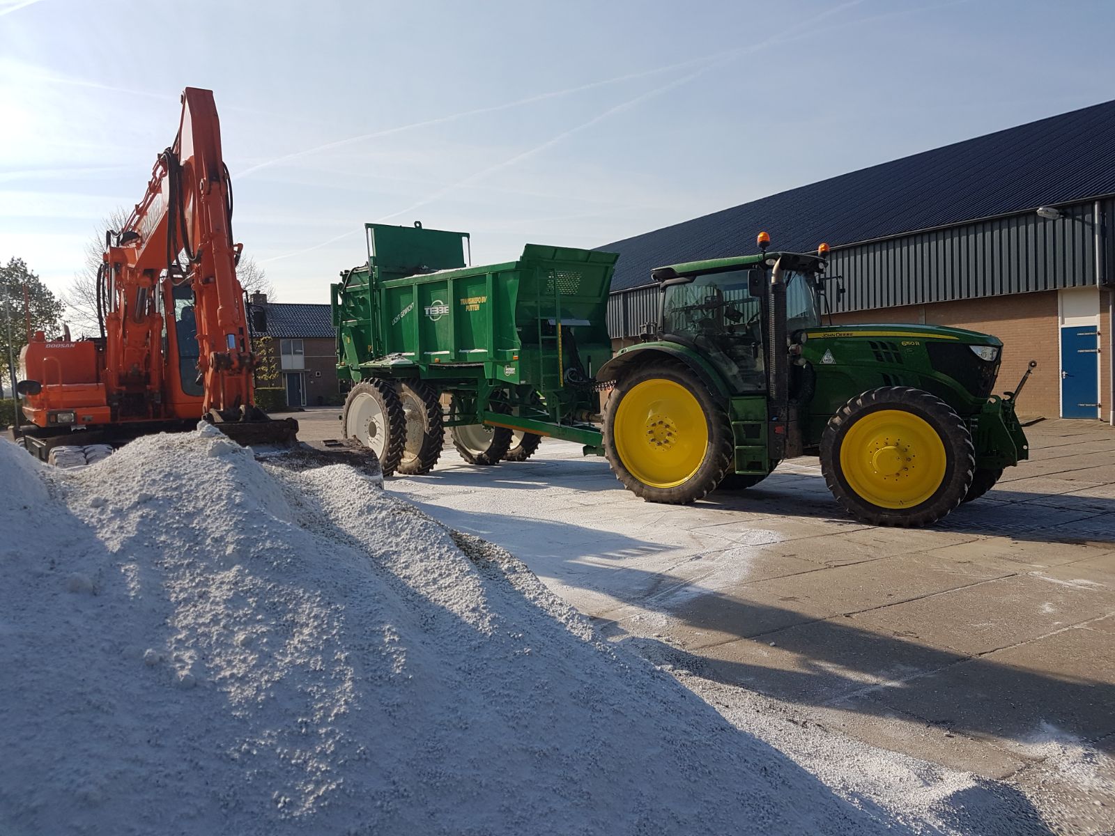 Oranje kraan en een John Deere trekker met verspreider naast een witte hoop op het erf