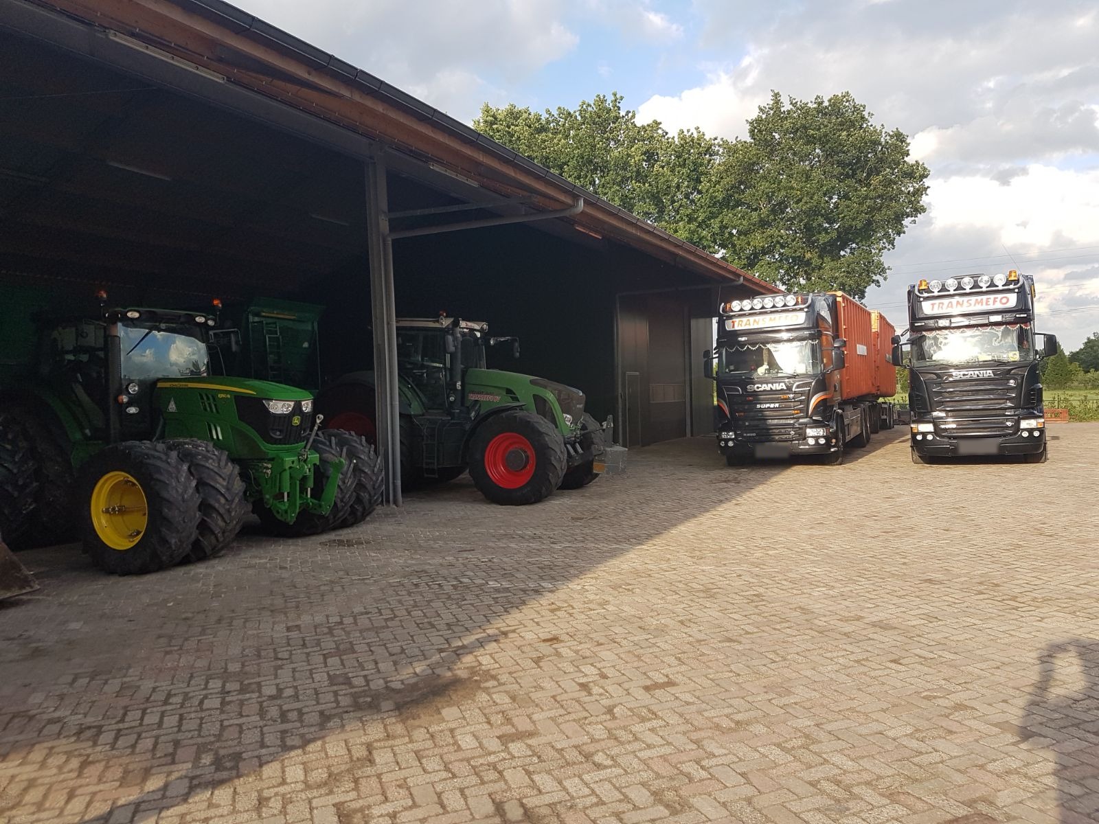 Trekkers in een open schuur en twee Scania-vrachtwagens met oranje containers op het klinkererf